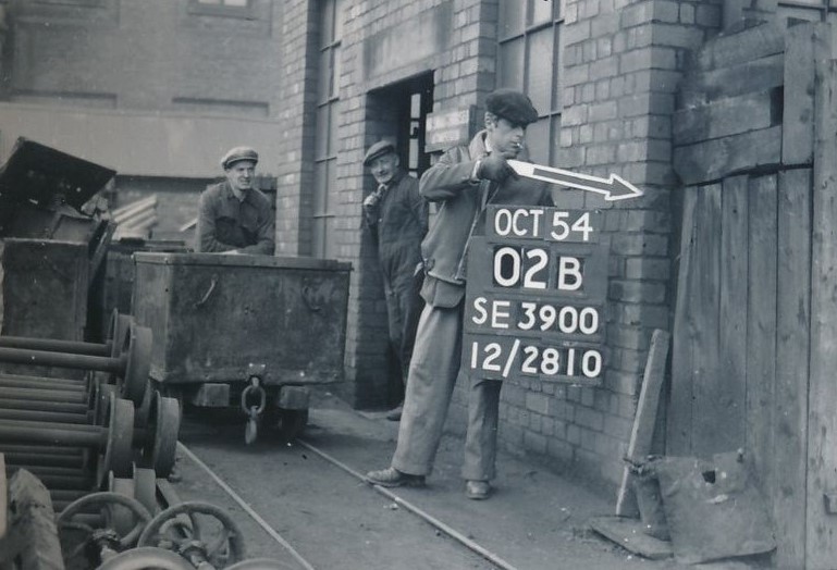 alt="work men in caps one with sign board and arrow next to an old building" 