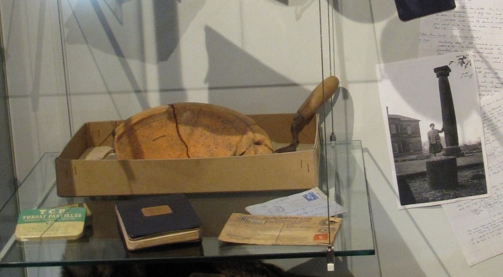 alt="museum display trowel and clay bowl in cardboard box with notebook and postcards in front"