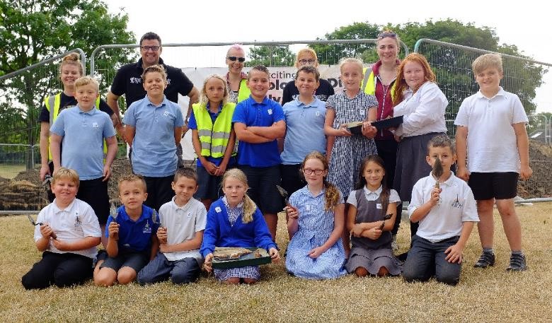 group of school children in front of trench holding trowels