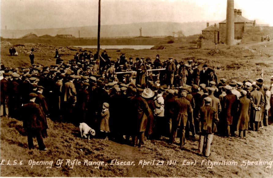 crowd in hats listening to speech with industrial buildings behind
