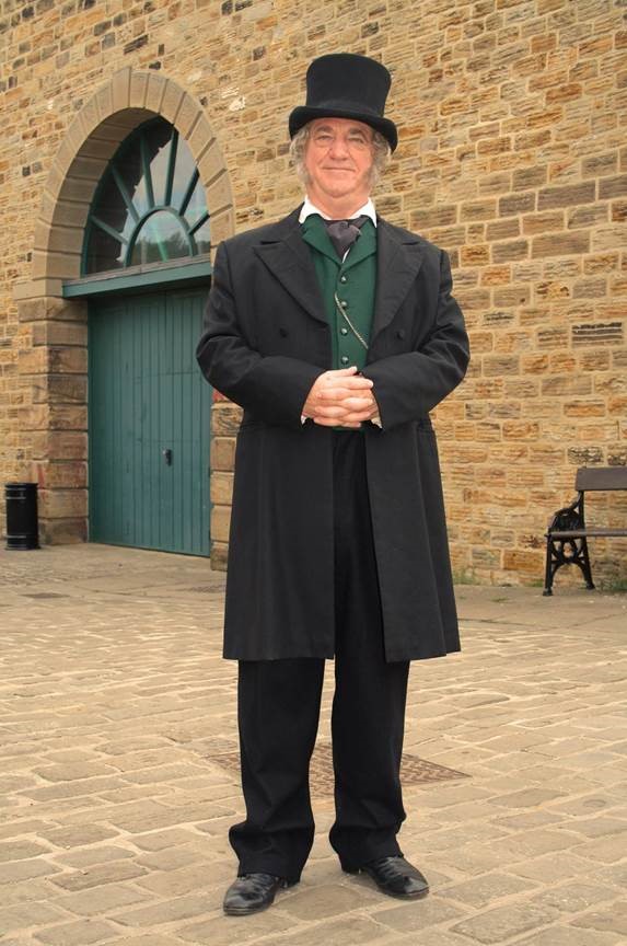 smiling man in top hat dark green waistcoat and long black coat in front of stone building