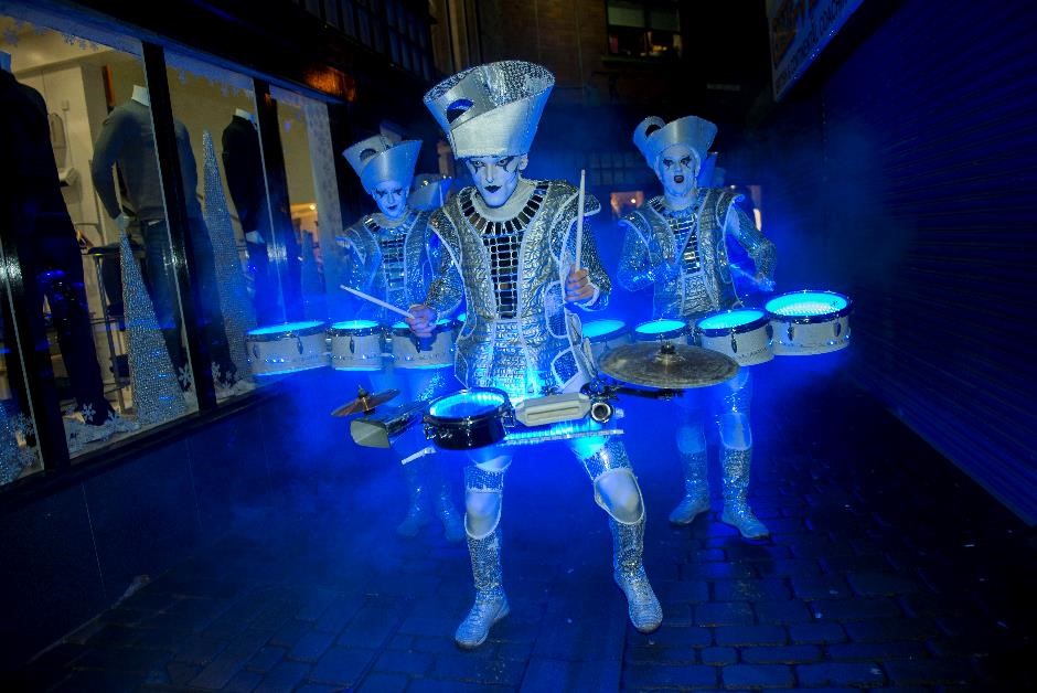 Three drummers in silver costumes with hats and facepaint glowing blue in darkened street
