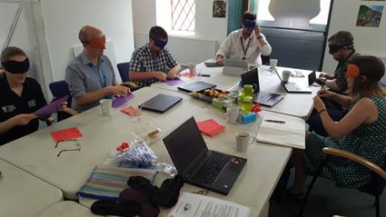 staff sat at a desk folding and making paper aeroplanes while wearing eye masks
