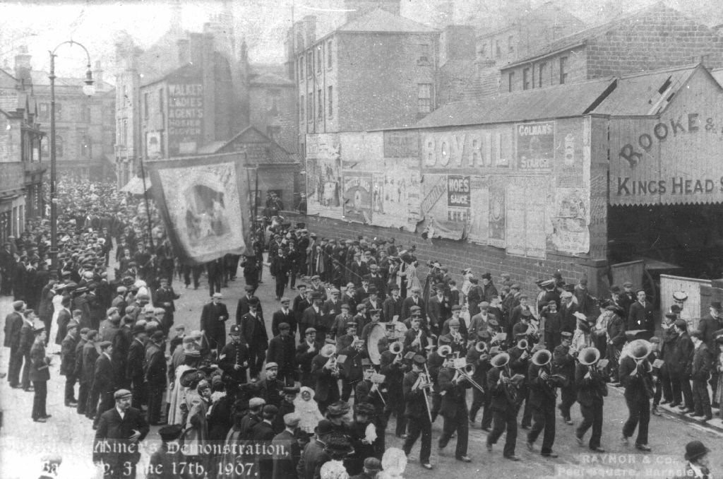 Black and white photograph people marching with brass band at the front and large square banner