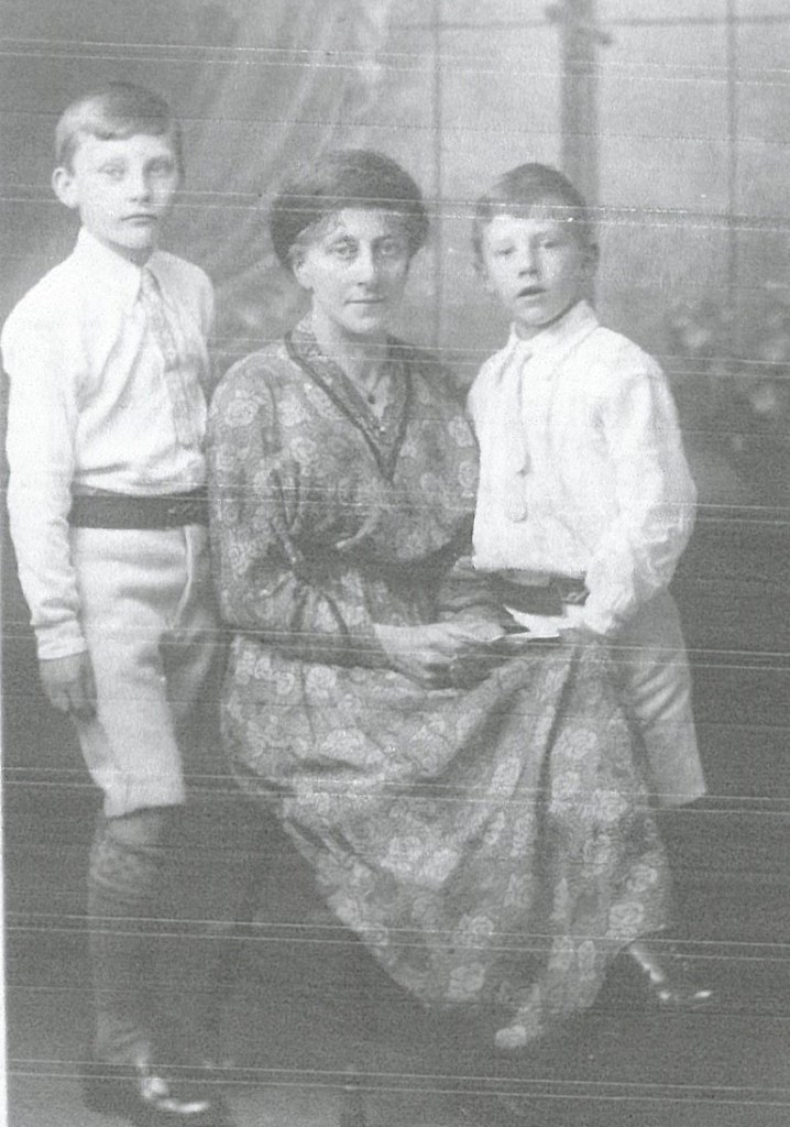 Black and white photo of lady sitting with two young boys to either side of her. She is in a long floral dress.