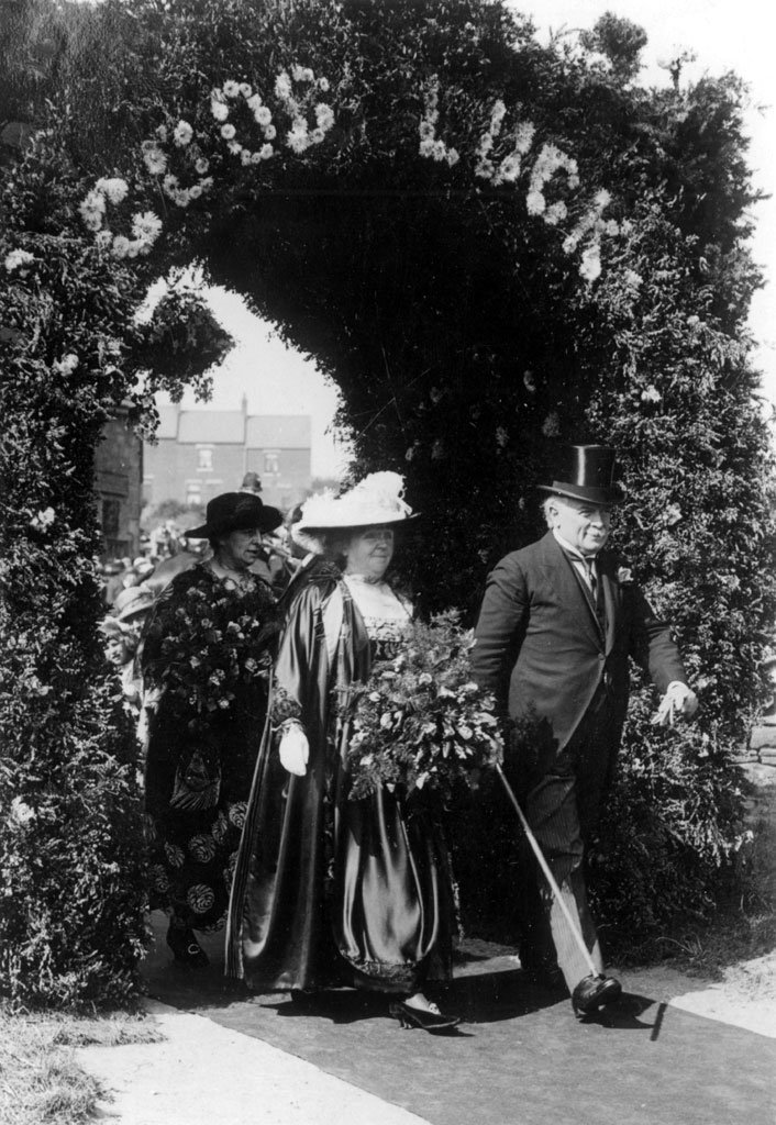 Mr and Mrs Lloyd George walking through an arch with 'Good Luck' written above. Both are wearing hats and Mrs Lloyd George is carrying a large bunch of flowers.