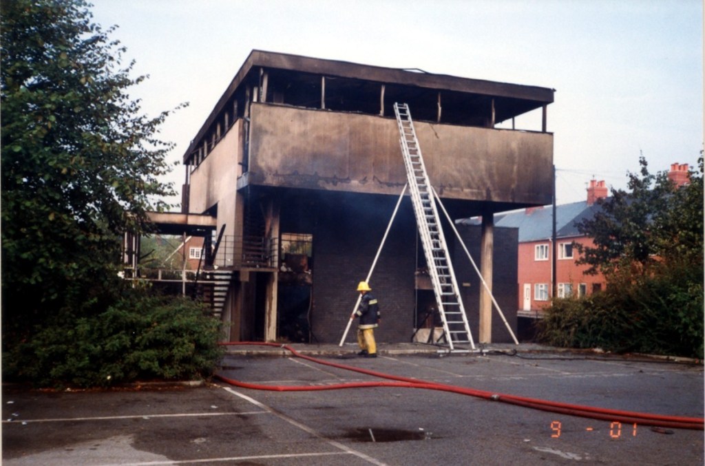 Goldthorpe Library after fire. Outside of building with blackened walls. Firefighter standing next to the building with ladder to first floor and fire hoses on the ground. 