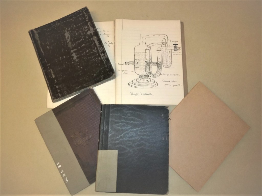Five exercise books on a table. One is open and shows a sketch of a scientific experiment (where a liquid is being heated and turned to gas).