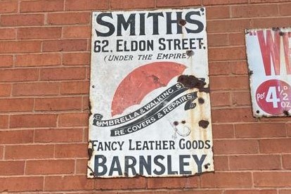 White enamel sign on a brick wall that says Smiths 62, Eldon Street (under the Empire) umbrella and walking stick re-covers and repairs, fancy leather goods, Barnsley. It has a picture of an umbrella in the middle.