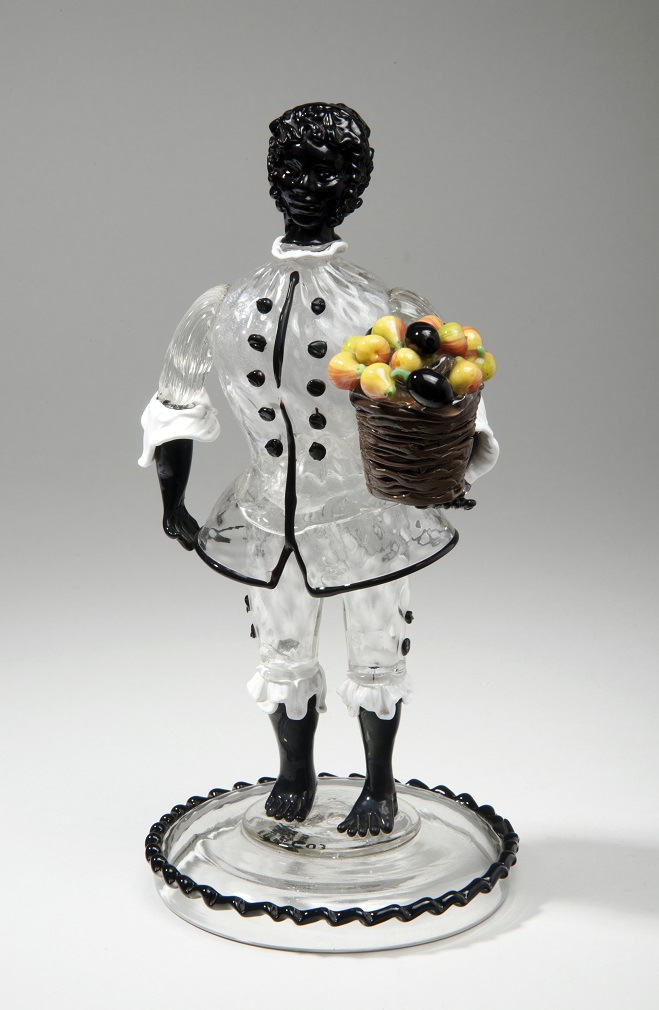 The figure's head, hands and feet are made from black glass and he has a clear glass tunic. He is holding a basket of fruit in his left hand.