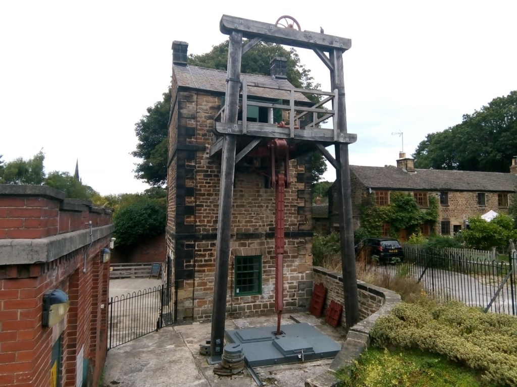 Photograph of the Elsecar Newcomen Engine as it is today. A tall, narrow stone building with a slate roof. It has a wooden frame in front of it and a long metal rod that stretched from the top floor of the building down into the ground below. There is a row of historic cottages to the right of the engine and a low curving stone wall in front.