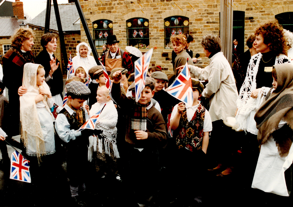 Group of adults and school children in Victorian costume waving union jack flags at the Elsecar Heritage Centre. Two little boys are staring straight at the camera and holding their flags in the air.