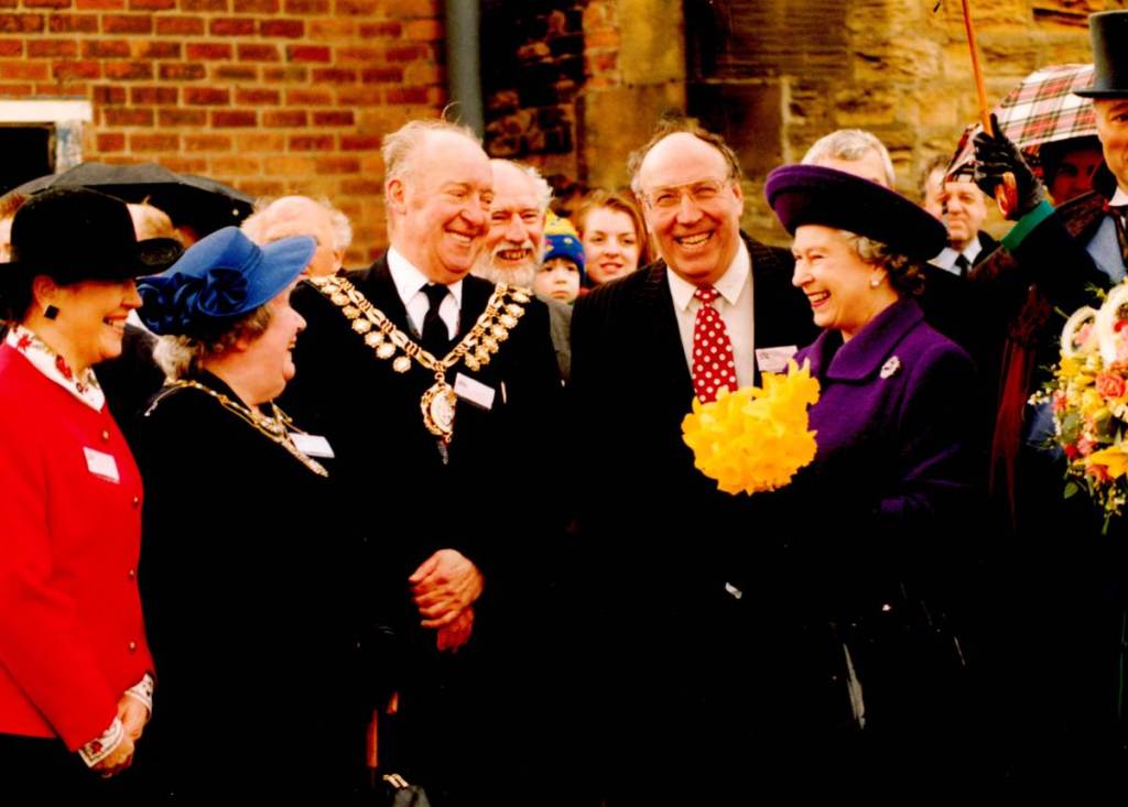 Group of smartly dressed people all smiling and laughing in front of one of the buildings in the Elsecar Heritage Centre. The group included the mayor of Barnsley and Queen Elizabeth II. The Queen is wearing a purple coat and hat and carrying a bunch of bright yellow daffodils.