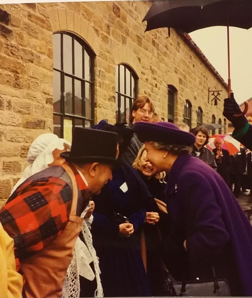 Queen Elizabeth at the opening of the Elsecar Heritage Centre. There is a crowd of people waiting and the Queen is talking to a man in a top hat at the front of the crowd. they are both smiling.
