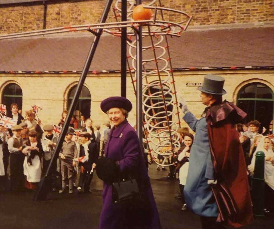 Queen Elizabeth walking across the courtyard at Elsecar Heritage Centre in front of a crowd of school children. She is looking at the camera and smiling. 