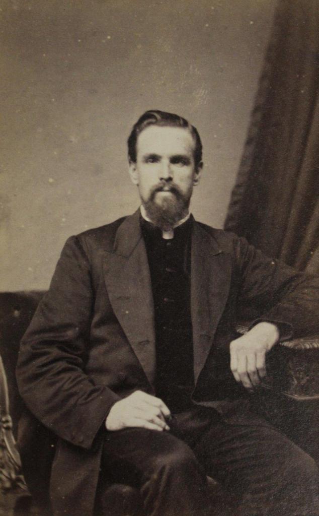 A vicar, his hair is pushed back, he is seated and looking towards the camera