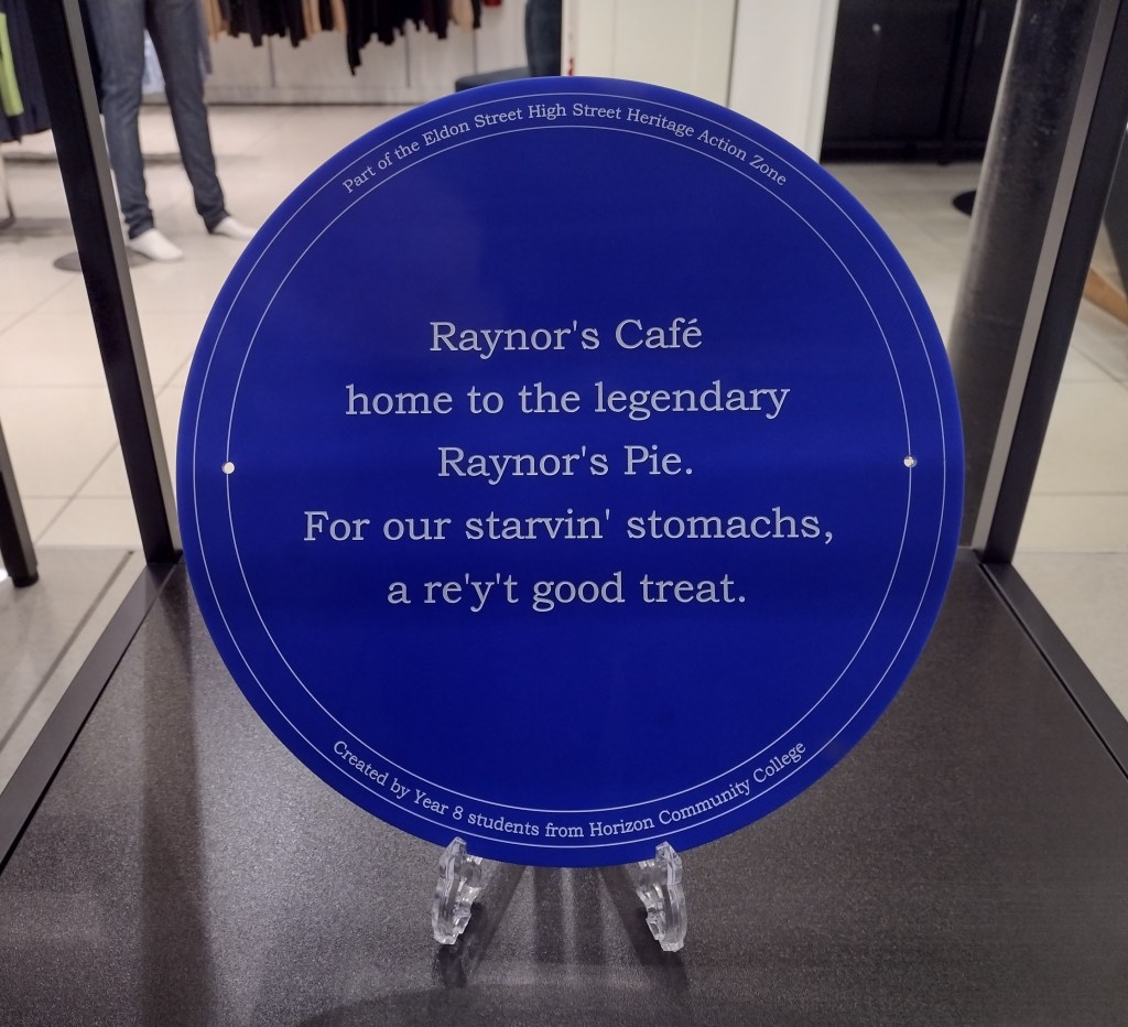 Blue plaque in a shop window. The text says 'Raynor's Cafe home to the legendary Raynor's Pie. For our starvin' stomachs, a re'y't good treat.' Text underneath says created by year 8 students from Horizon College.