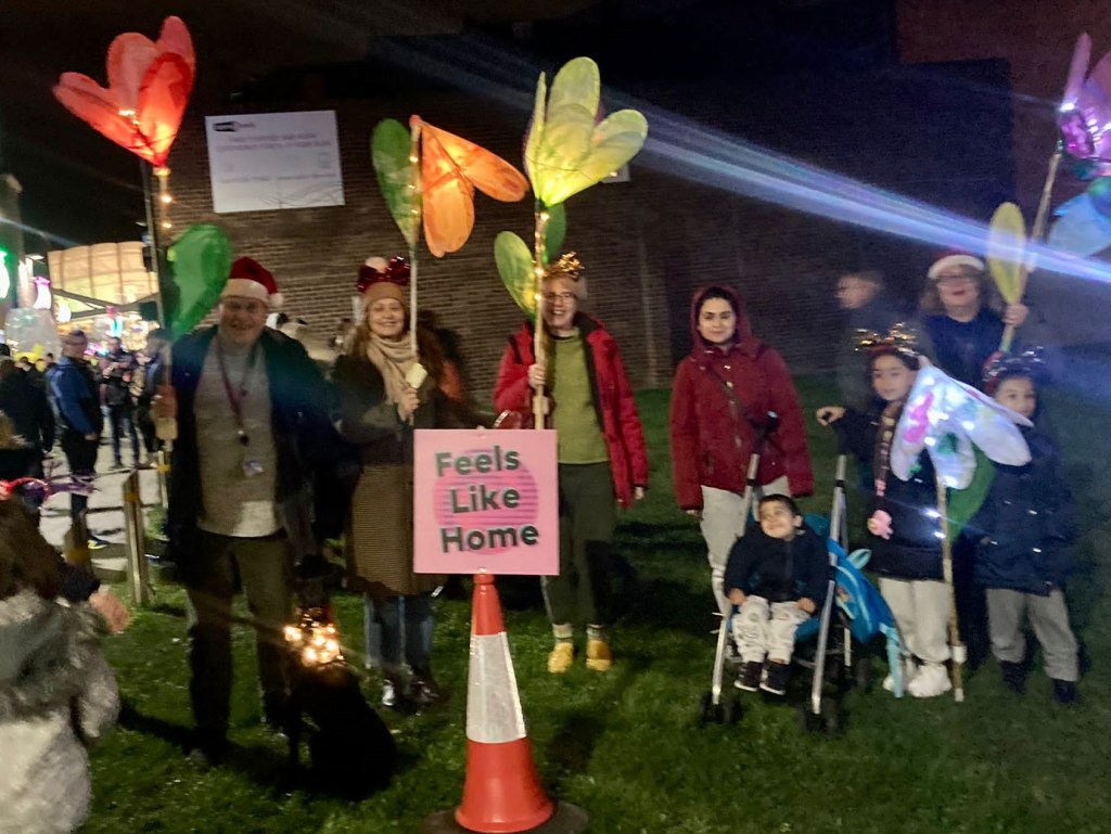 Group of people (adults and children) holding large colourful, illuminated flowers. There is a dog with the group with led lights around its collar. A sign in front of the group says 'Feels Like Home'