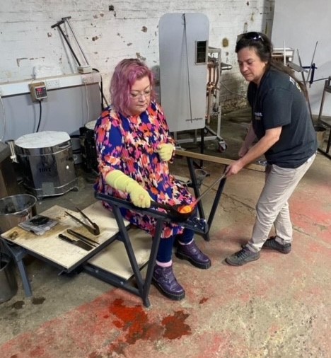 Volunteer having a go at glass blowing at Elsecar Heritage Centre. They are sitting in a metal chair with heat proof gloves and safety goggles on, holding a small black pan full of molten glass. The workshop leader is standing next to them with a glass blowing tube in their hands. They are putting the tube into the molten glass.