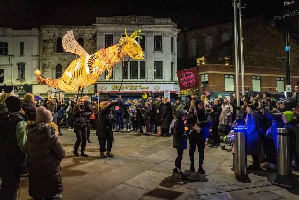 Eldon Beast puppet surrounded by crowds of people on Eldon Street. Lots of people are taking photographs. The beast is a large illuminated puppet with a rabbit's head, a tiger's tail, slug horns and wings. It is held above head height on poles and operated by four people standing underneath. It looks like it is flying.