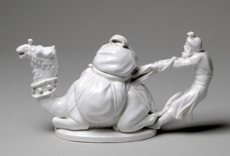 A ceramic teapot made from porcelain or bone china moulded in the form of a camel and its handler. The camel's head forms the spout and the handler forms the handle. The lid is in the form of a package on the camel's back.