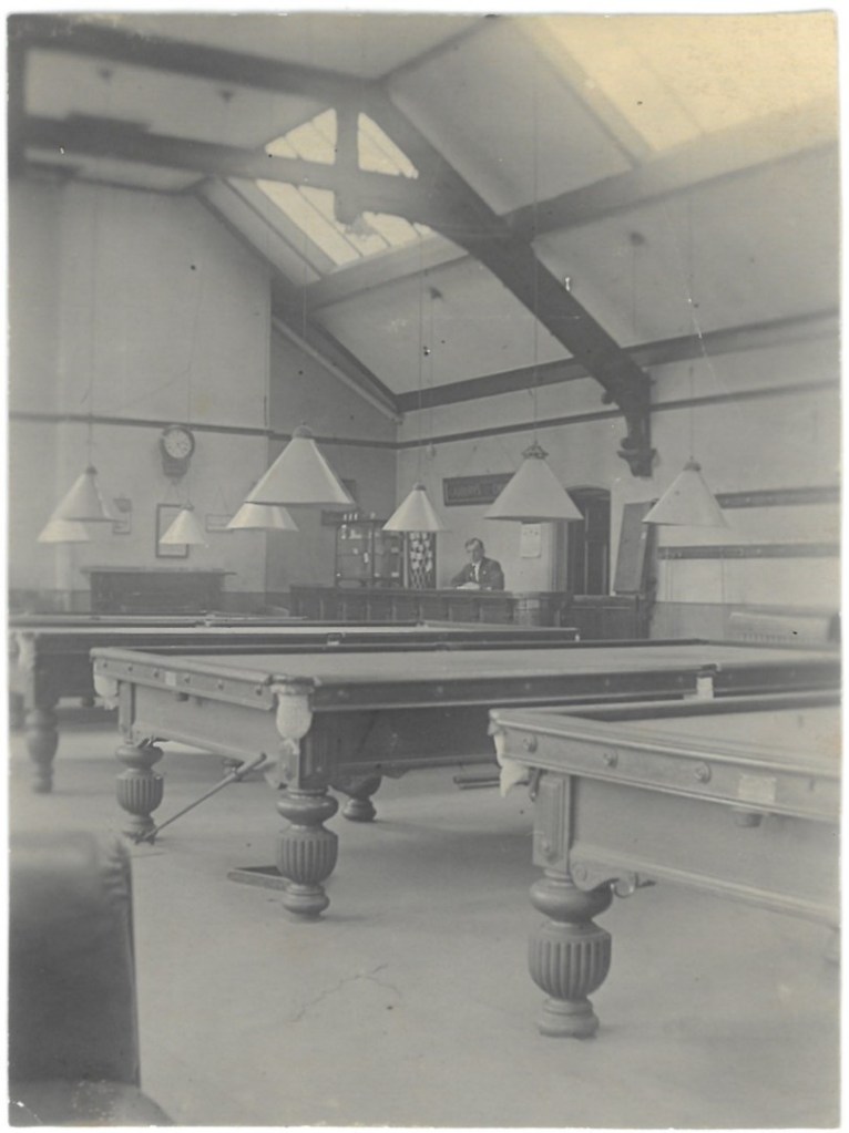 Photograph of the billiard room on the top floor the YMCA building in the early 1900s. There are three billiard/pool tables in shot, with hanging lights above. In the corner of the room in a man sitting behind a desk. There are skylights in the roof above and exposed decorative wooden beams.