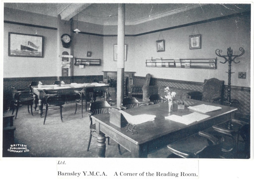Photograph entitled ' Barnsley YMCA a corner of the reading room' It shows a room with tables and chairs, with a clock and racks for newspapers on the wall. Other features include a painting of a steam ship and a hat stand, and a corner fireplace. The decoration looks very smart, with a wooden dado rail running along the walls.