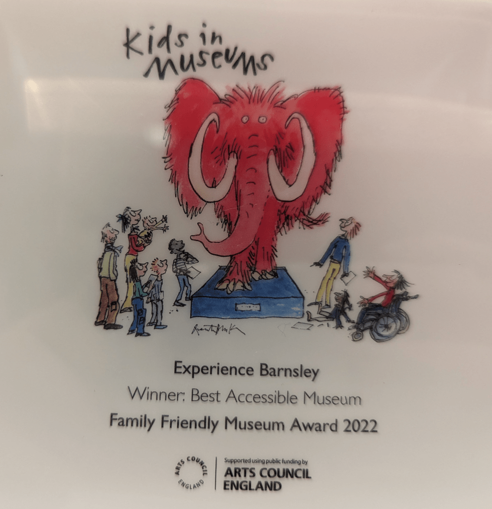 The Experience Barnsley Kids In Museum Family Friendly award featuring the kids in museum logo, a pink elephant on a stand with lots of people stood around looking 