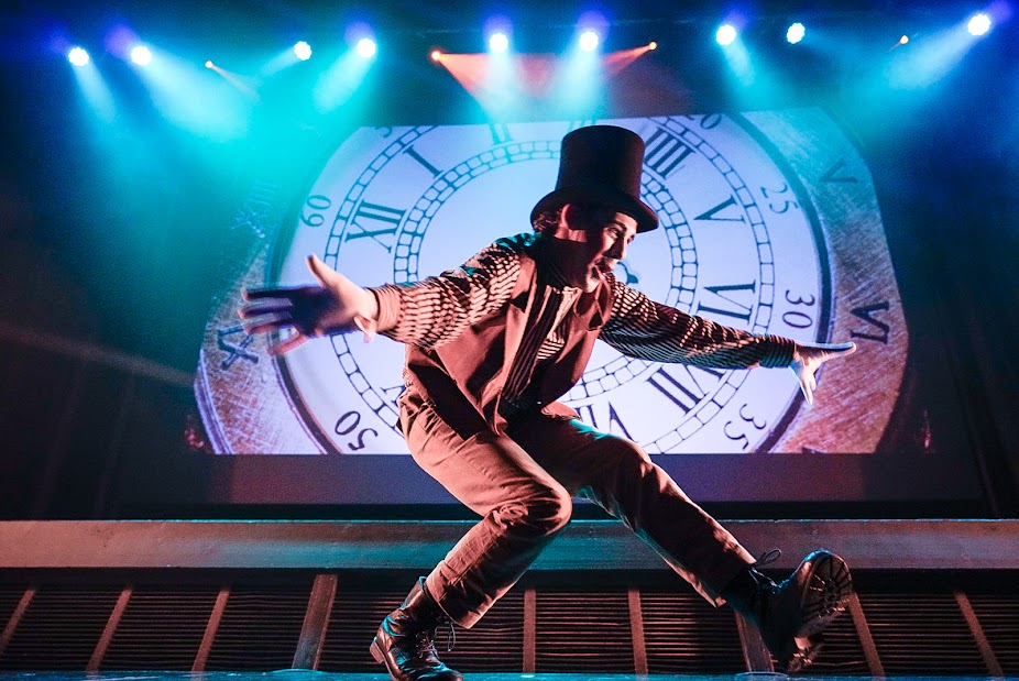 Dancer dressed in a top hat, shirt, trousers and waistcoat with open arms on stage in front of a large projected clock