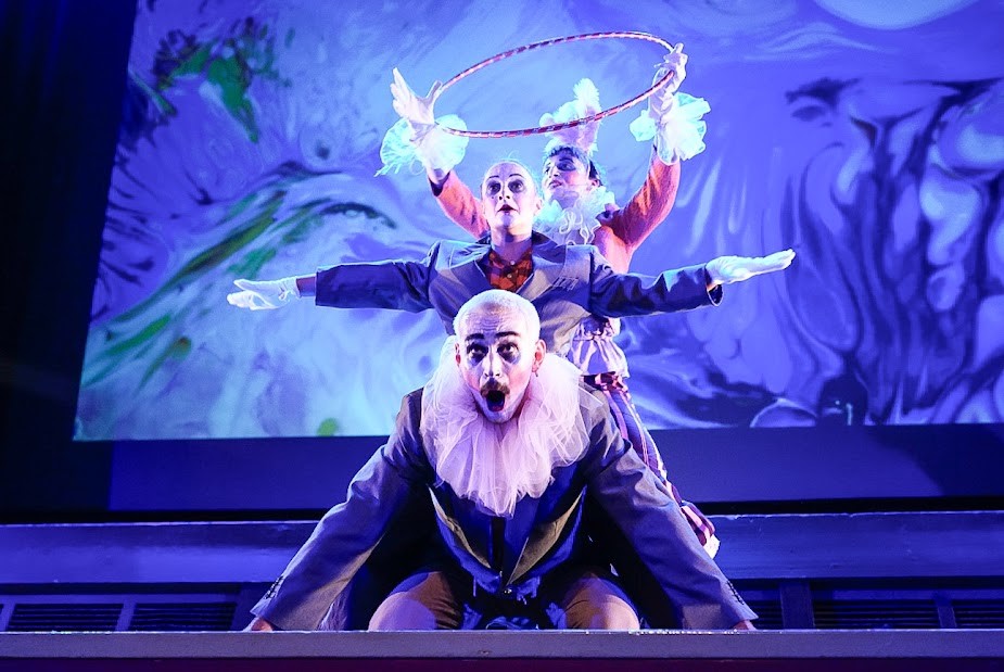 Three dancers dressed as clowns in formation on stage depicting circus acrobats. The first dancer is kneeling down, the second dancer (behind) is standing with their arms outstretched and the the third dancer (at the back) is holding up a large hoop