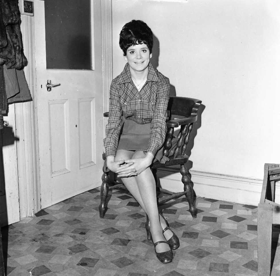 A girl with a beehive hair-do sitting on a chair and sixties clothes (skirt and checked shirt) sitting on a chair