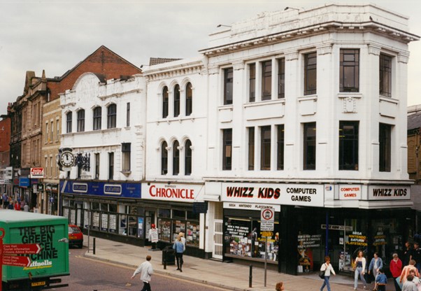 Colour photograph of 32-38 Eldon Street. The buildings have all been painted white. 34 has a large sign that says Barnsley Chronicle above the shop front and 36-38 has a sign saying Whizz Kids computer games.