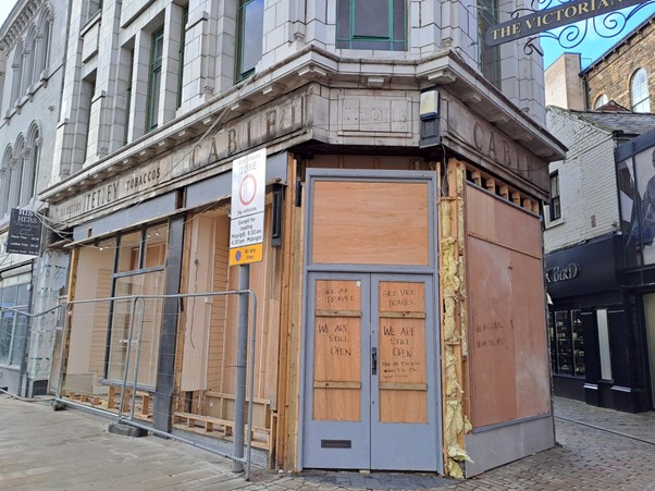 Colour photograph of the shopfront at 36-38 Eldon Street during restoration works. the shop windows and door have all been boarded and there are historic ghost signs visible above. One reads Tetley cigarettes tobaccos, and one reads Cable shoes Cable. There is a written sign on the boarding of the door that says ;We Are Open'