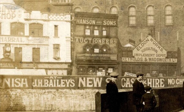 Black and white photo showing 32, 34 and 36 Eldon Street. 24 has large painted signs between each floor of the building that say Hanson and Sons Auctioneers Philanthropic Friendly Collecting Society. Number 36 is a single storey building with a large sign that reads 'Eldon Works, agricultural implement depot, Reynolds and Wadsworth Ltd.' There is also painted signage along the side of the road that says 'J H Baileys New St, Cash or Hire' with two men and a boy in flat caps standing in front of the sign. One is looking towards the camera.