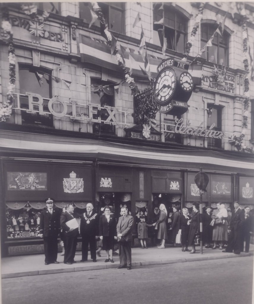 black and white photograph of the Ring Shop frontage covered in bunting and royal banners in readiness for Queen Elizabeth's coronation. A group of people including the mayor of Barnsley (with his civic chain) are standing outside the shop, all dressed smartly and looking like they are waiting for somebody.