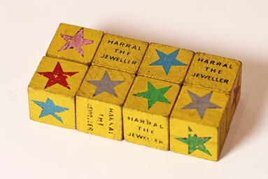 8 yellow dice with a painted star in different colours on each face. On one side of four of the dice are the words 'Harral the Jeweller'