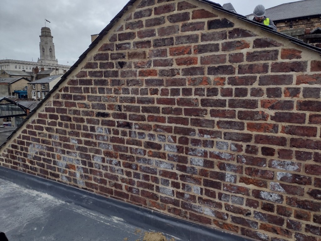 Top of a brick gable wall that has recently been repointed. The faded outline of white painted letters 'CHARDS' are visible on the paintwork. They have been painted in an arch, and only the top of the arch is now visible. A person in a high vis and hard hat is visible working on the roof behind. The tower of Barnsley Town Hall is also visible in the background