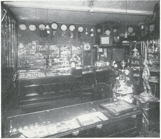 Shop interior with wood and glass counter top display cases and lots of clocks hanging from the ceiling. There is a decorative lamp next to the counter with the figure of a young person in a hat and a light fitting extending our of the top of the figurine.