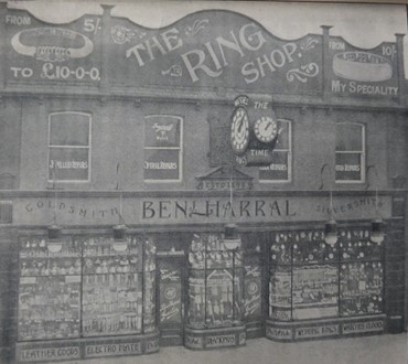 Black and white photo of the pre-1904 Ring Shop frontage. It is a two-storey building with a large decorative clock projecting into the street at first floor level above a tiled fascia sign that says Benj Harral Goldsmith Silversmith. There is a large sign board above the first floor windows that says the Ring Shop. there are a lot of clocks, watches and other goods displayed in the ground floor windows. The windows on the first floor match those shown on the previous architects plan.