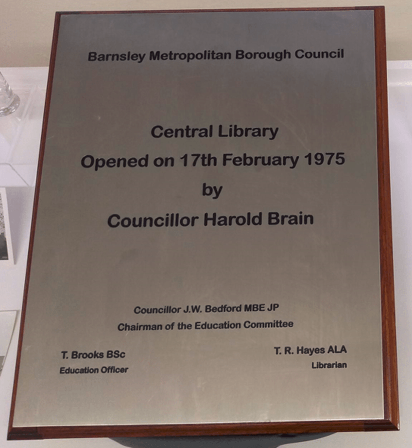 A plaque with a wooden frame which reads: Barnsley Metropolitan Borough Council, Central Library opened on 17th February 1975 by councillor Harold Brain, Councillor J.W. Bedford MBE JP Chairman of the Education Committee, T. Brooks BSc Education Officer, T.R. Hayes ALA Librarian
