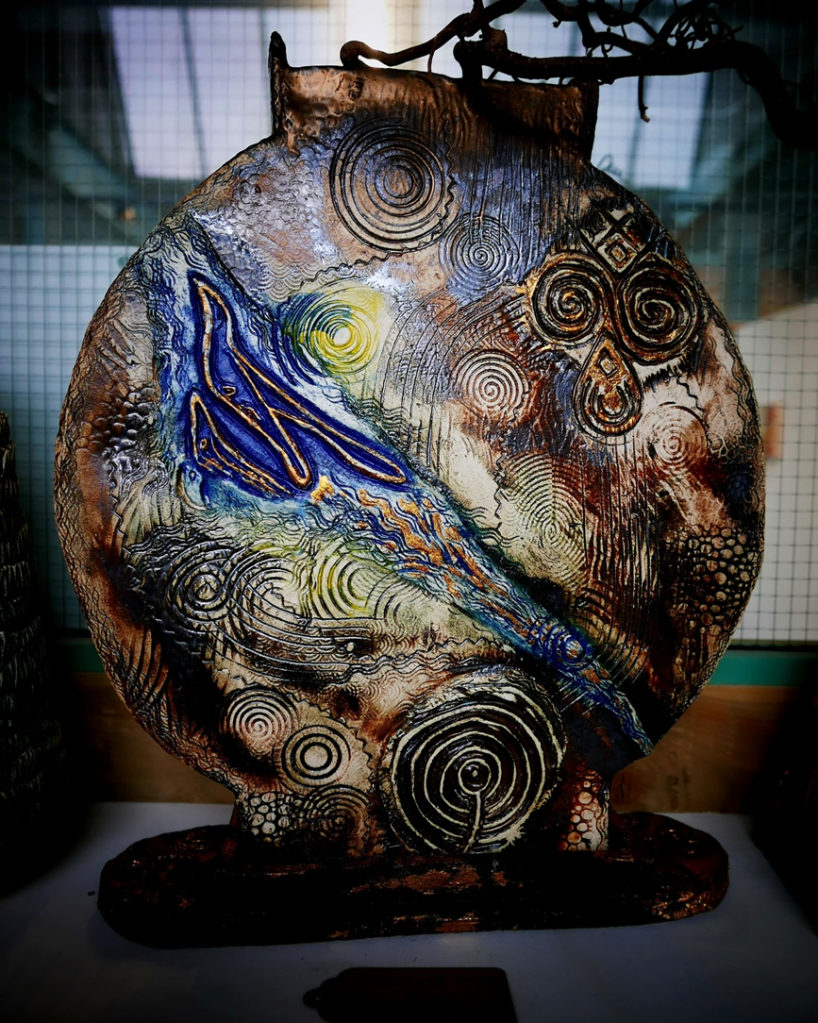  A circular ceramic artwork with abstract patterns, including swirls and a blue wave-like design, displayed indoors against a blurred background.
