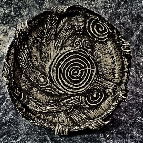 A round, stone-like object is intricately carved with spiral and circular patterns. The texture appears rough and weathered, giving it an ancient and rustic appearance. The background is a dark, marbled surface, enhancing the contrast of the carvings.