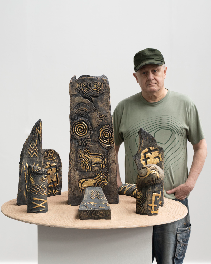 A man wearing a green cap and a green patterned t-shirt stands next to a round table displaying several abstract, carved sculptures with intricate patterns. The background is plain white.
