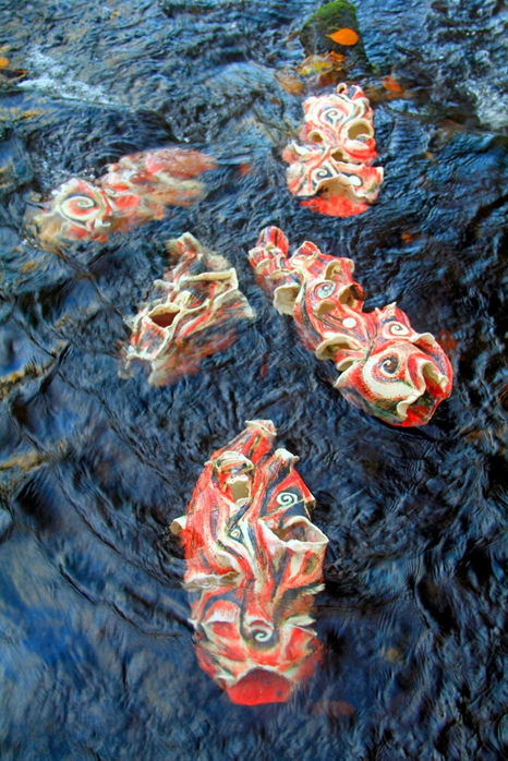 Five brightly colored, intricately designed ceramic masks, painted primarily in red, white, and black, float on the surface of dark, flowing water. The masks feature swirling patterns and exaggerated facial features, creating a striking contrast with the water.