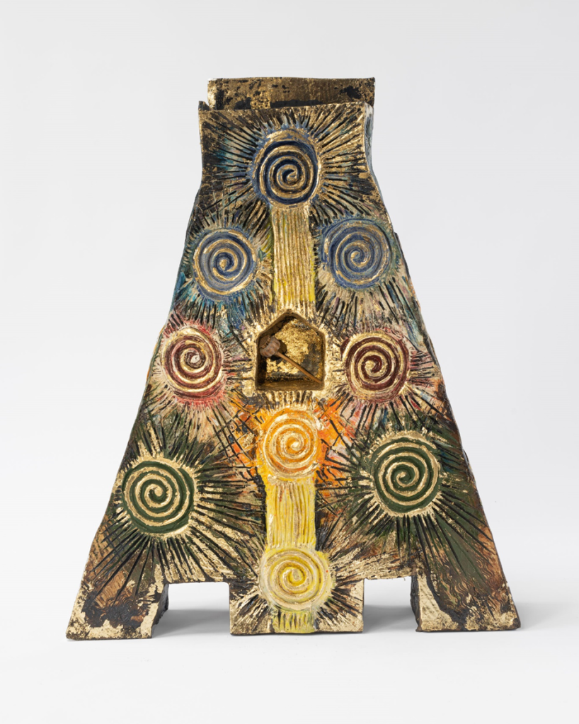 A triangular-shaped ceramic sculpture features colorful swirls and radiating lines in blue, green, red, yellow, and gold. The textured patterns converge at a small central cavity, which contains a raised, gold-colored square. The surface has a rustic, aged appearance.