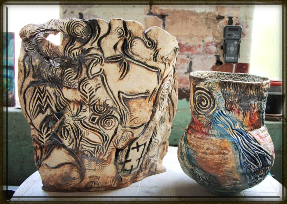 A photo shows two intricately decorated pottery pieces. The larger piece has abstract and tribal-like carvings, while the smaller, rounder vessel features colorful patterns, including a prominent blue and green motif. Both rest on a white table in a rustic setting.