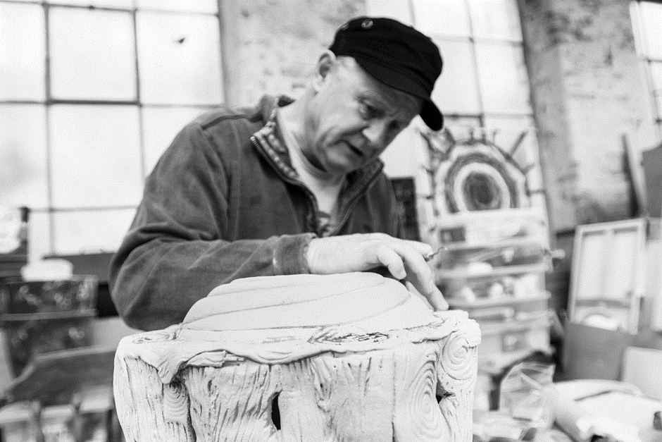 A man wearing a cap and a jacket concentrates on carving a sculpture inside a workshop. The background shows a large window letting in natural light and various tools and materials scattered around.