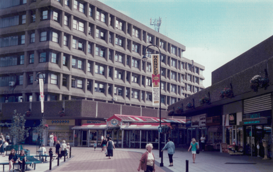 Central Offices in background with entrance to open market below and Metropolitan Centre shops to the right. People walking along the street or sitting on benches.