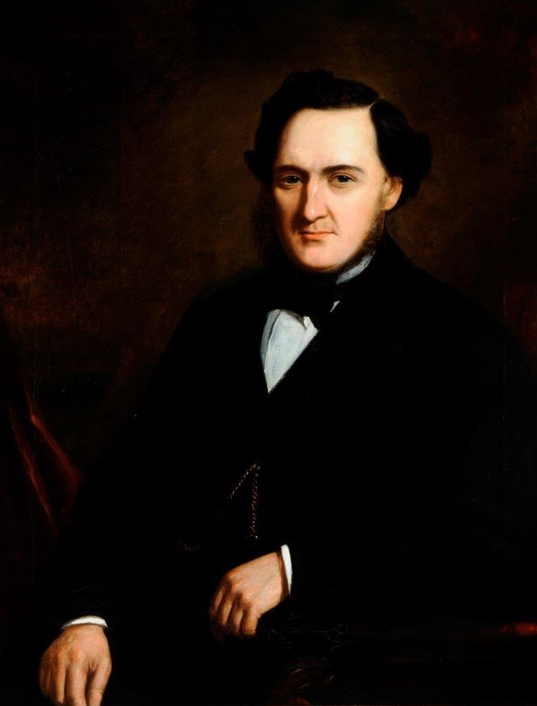A portrait of a man in formal 19th-century attire. He has dark hair with sideburns, and he is wearing a black coat with a white shirt and a black bow tie. The background is dark, highlighting his serious expression and composed demeanor.