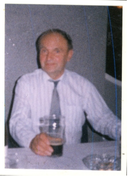 A man with a receding hairline in a white dress shirt and gray tie is sitting at a table, holding a glass of dark beer. The image is slightly blurry. A white wall and a partial view of another person are visible in the background.